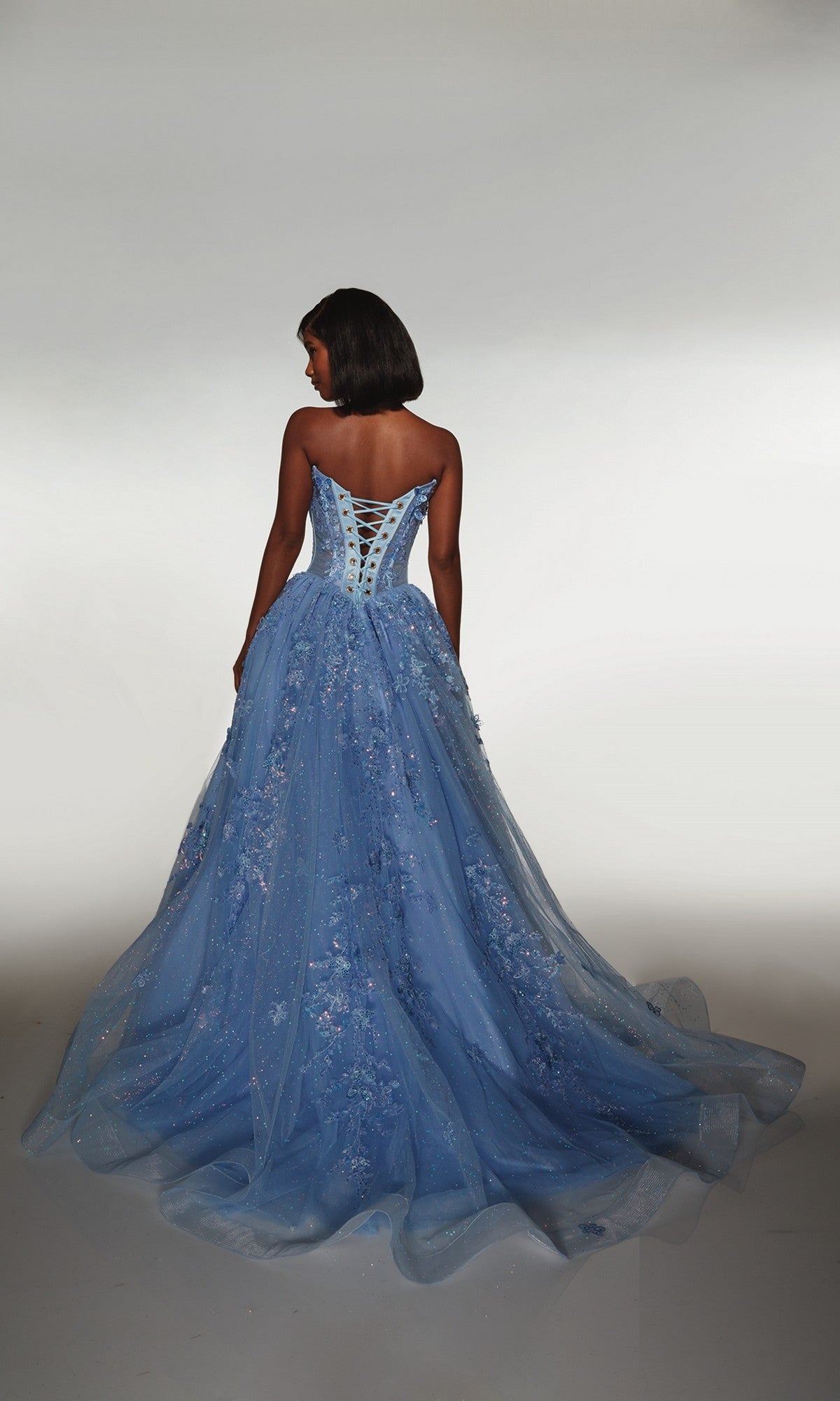 A woman in the Alyce 62163 Long Prom Dress, a strapless blue gown with an embellished corset bodice and floral details, stands facing away against a plain light background.